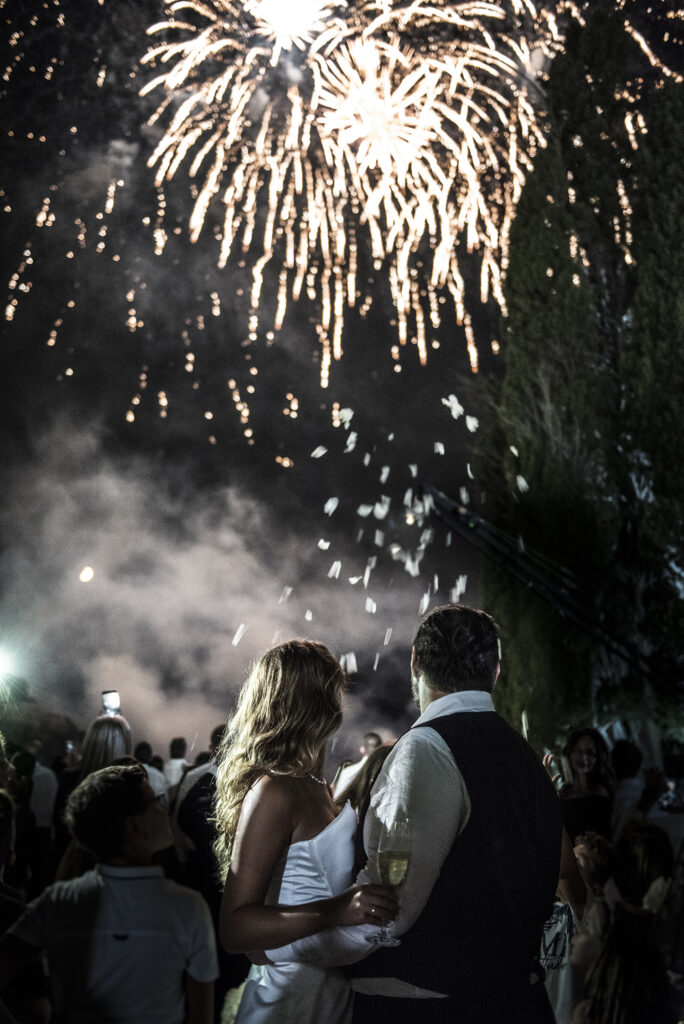 A bride & groom standing together with their backs facing the camera as they watch the fireworks above them
