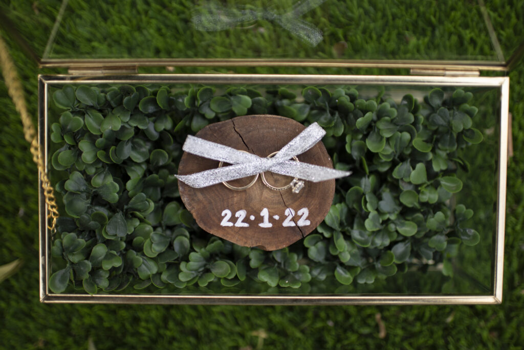 Glass box with wedding rings inside, tied with a silver ribbon on a small piece of wood with the date 22/1/22 written on it. Sitting on a green lawn with greenery inside the box as well